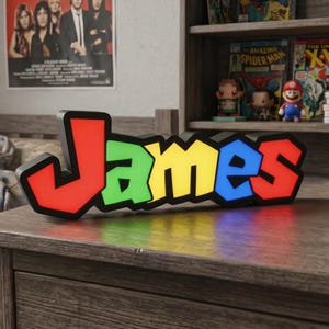 May include: A custom 3D-printed sign displaying the name James in a bold, blocky font. The letters are individually coloured in red, green, yellow, and blue, featuring a thick black outline that gives the sign a distinct, layered appearance.