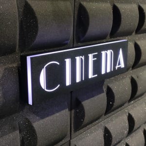 Pode incluir: Uma placa preta com letras brancas que lê "CINEMA" está montada em uma parede texturizada preta.