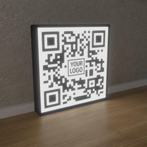 May include: A lit-up QR code sign with a black frame and a white QR code pattern. The center of the code has the text "YOUR LOGO". The sign is leaning against a gray wall on a wooden floor.