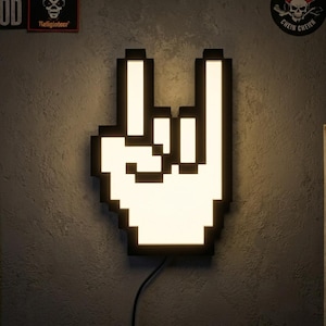 Può includere: Una lampada da parete a LED a forma di mano pixelata che fa il segno del rock and roll. Il design presenta una cornice nera con un centro illuminato bianco brillante, creando una silhouette grafica ad alto contrasto sulla superficie murale.