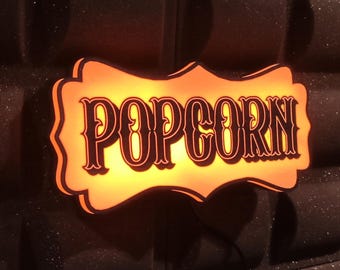 May include: A bright orange sign with a black outline and the word "POPCORN" in a decorative font.