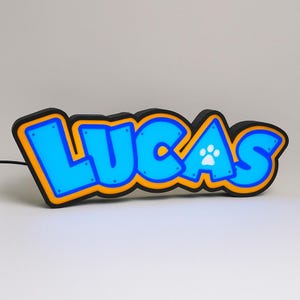 Pode incluir: Letreiro luminoso LED azul com o nome "LUCAS" em uma fonte estilo grafite. As letras são contornadas em laranja e preto, com uma pegada na letra "S". Projetado como um item decorativo.