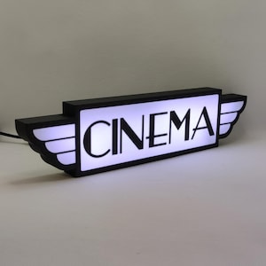 Pode incluir: Placa iluminada em preto e branco com a palavra "CINEMA" em uma fonte retrô, com asas de cada lado.