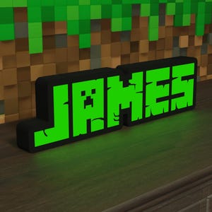 Może przedstawiać: Czarny i zielony podświetlany znak z napisem "JAMES" w pikselowanej czcionce. Znak znajduje się na drewnianej powierzchni z pikselowanym tłem. Zielone litery świecą jasno, tworząc uderzający kontrast z czernią.