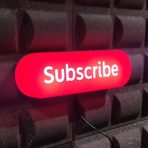 Op de afbeelding: Een rood, rechthoekig bord met afgeronde hoeken waarop in witte letters "Subscribe" staat.