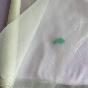 May include: A roll of sheer, off-white fabric partially unrolled, revealing a translucent texture. A small, bright green feather rests on the fabric. The fabric appears to be lightweight and suitable for crafts or decorative purposes.
