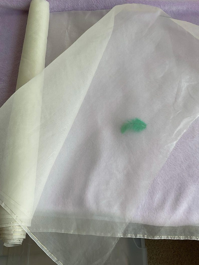 May include: A roll of sheer, off-white fabric partially unrolled, revealing a translucent texture. A small, bright green feather rests on the fabric. The fabric appears to be lightweight and suitable for crafts or decorative purposes.