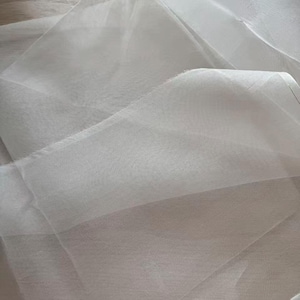 May include: A close-up shot of a sheer, white fabric with a delicate, flowing texture. The fabric is folded and layered, creating shadows and highlights that emphasize its translucence. The material appears soft and lightweight.