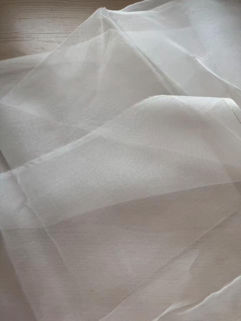 May include: A close-up shot of a sheer, white fabric with a delicate, flowing texture. The fabric is folded and layered, creating shadows and highlights that emphasize its translucence. The material appears soft and lightweight.