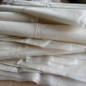 May include: A stack of off-white fabric rolls and folded sheets. The fabric appears to be lightweight and possibly sheer, with a soft, delicate texture. The image shows a close-up of the fabric, highlighting its smooth surface and the way it drapes.