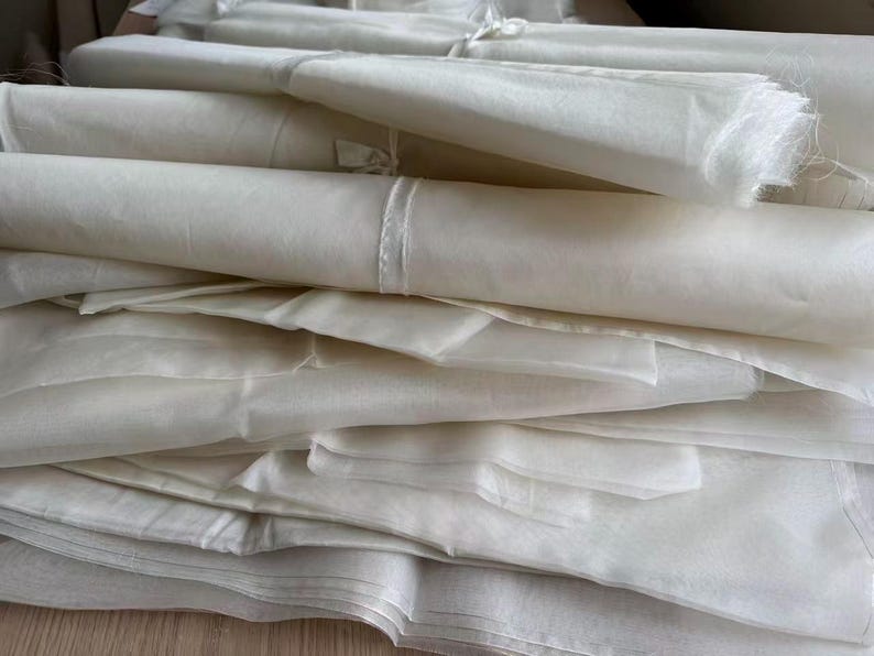 May include: A stack of off-white fabric rolls and folded sheets. The fabric appears to be lightweight and possibly sheer, with a soft, delicate texture. The image shows a close-up of the fabric, highlighting its smooth surface and the way it drapes.
