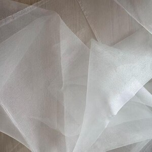 May include: Close-up of white sheer fabric, possibly tulle or organza, with a delicate, flowing texture. The fabric is layered and folded, creating shadows and depth. The material appears lightweight and translucent, ideal for crafting or decorative purposes.