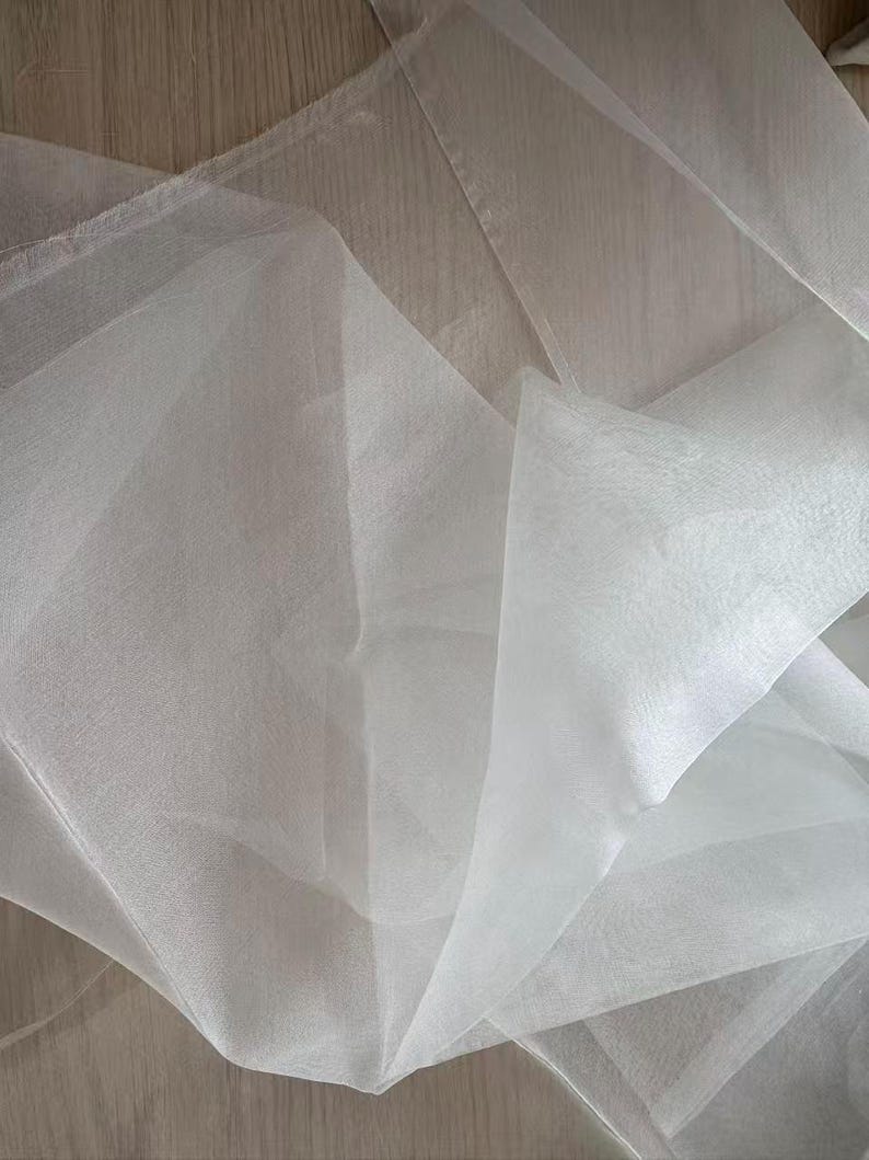 May include: Close-up of white sheer fabric, possibly tulle or organza, with a delicate, flowing texture. The fabric is layered and folded, creating shadows and depth. The material appears lightweight and translucent, ideal for crafting or decorative purposes.