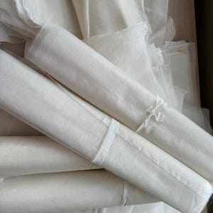 May include: A collection of white, sheer fabric rolls and folded sheets. The fabric appears lightweight and delicate, with a slightly translucent quality. Some rolls are secured with white ribbon. The overall impression is of soft, flowing material.
