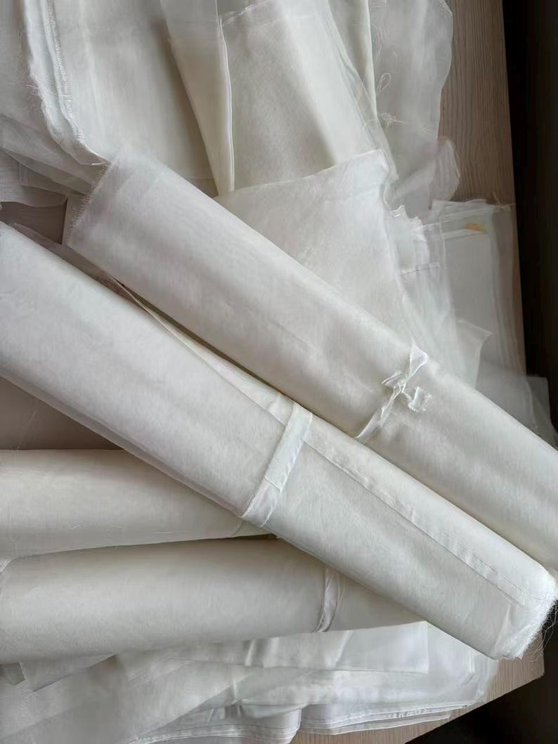 May include: A collection of white, sheer fabric rolls and folded sheets. The fabric appears lightweight and delicate, with a slightly translucent quality. Some rolls are secured with white ribbon. The overall impression is of soft, flowing material.