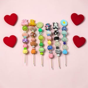 May include: Six decorative pens featuring colorful beaded designs with various toppers including a pink pig, yellow bear, green flower, cow, dog, and blue flower. Each pen is adorned with a mix of textured, patterned, and solid-colored beads.