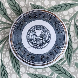 May include: A vintage white ceramic lid with a gold rim and black lettering. The lid reads "The Areca Nut Tooth Paste" and features a black and white logo of the Army & Navy Co-Operative Society Limited.