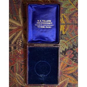 May include: An open, antique jewelry box with a deep blue velvet interior. The box is lined with a dark blue velvet and has the text "W.E. FELLOWS JEWELLER & SILVERSMITH" printed on the inside. The exterior is a dark brown color.