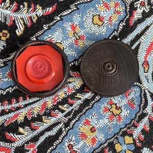 May include: A red wax seal with a crescent and wreath design inside a wooden octagonal box. The lid of the box is next to it. The seal is on a patterned blue, red, and yellow textile.