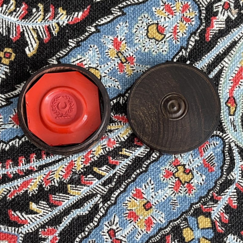 May include: A red wax seal with a crescent and wreath design inside a wooden octagonal box. The lid of the box is next to it. The seal is on a patterned blue, red, and yellow textile.