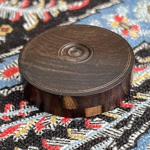 May include: A round, dark brown wooden box with a lid. The lid has a circular design in the center. The box is made of a dark wood with lighter wood accents. The box is sitting on a patterned surface.