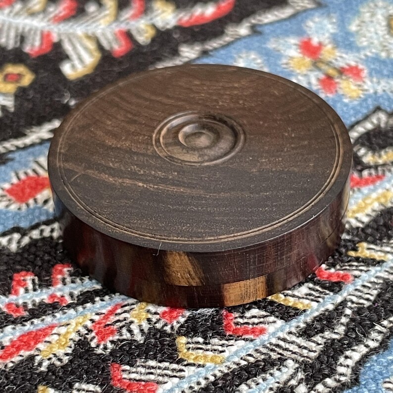 May include: A round, dark brown wooden box with a lid. The lid has a circular design in the center. The box is made of a dark wood with lighter wood accents. The box is sitting on a patterned surface.