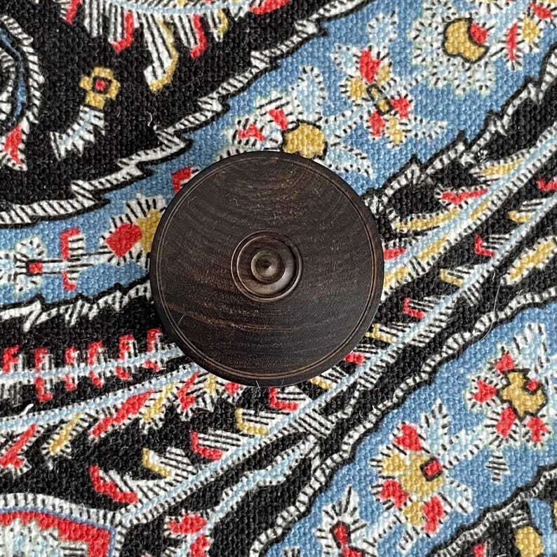 May include: A dark brown, circular wooden object with a central, concentric design. The object is set against a patterned background with blue, black, red, and yellow floral motifs. The wood grain is visible.