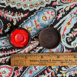 May include: A red wax seal with a crest design inside a dark wooden case, next to an empty wooden case. A wooden ruler with text is below the cases. The text on the ruler says "For Desk or Basket" and "Reliance Printing & Stationery Co. Inc."