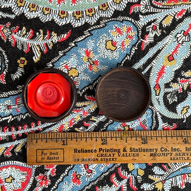May include: A red wax seal with a crest design inside a dark wooden case, next to an empty wooden case. A wooden ruler with text is below the cases. The text on the ruler says "For Desk or Basket" and "Reliance Printing & Stationery Co. Inc."