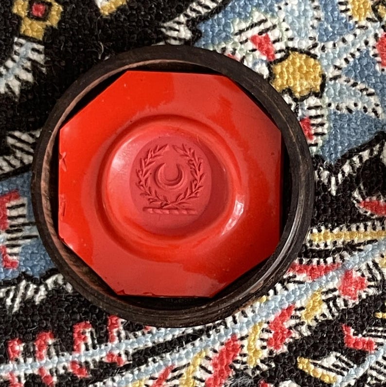 May include: A red wax seal with a crescent moon and wreath design, set within a dark wooden octagonal frame. The seal is a vibrant red, and the design is embossed. The background features a colorful patterned textile.