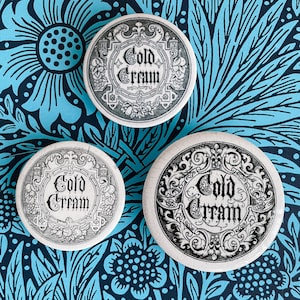 May include: Three white ceramic jar lids with black and white designs. Each lid features a decorative border and the words "Cold Cream" in a stylized font.