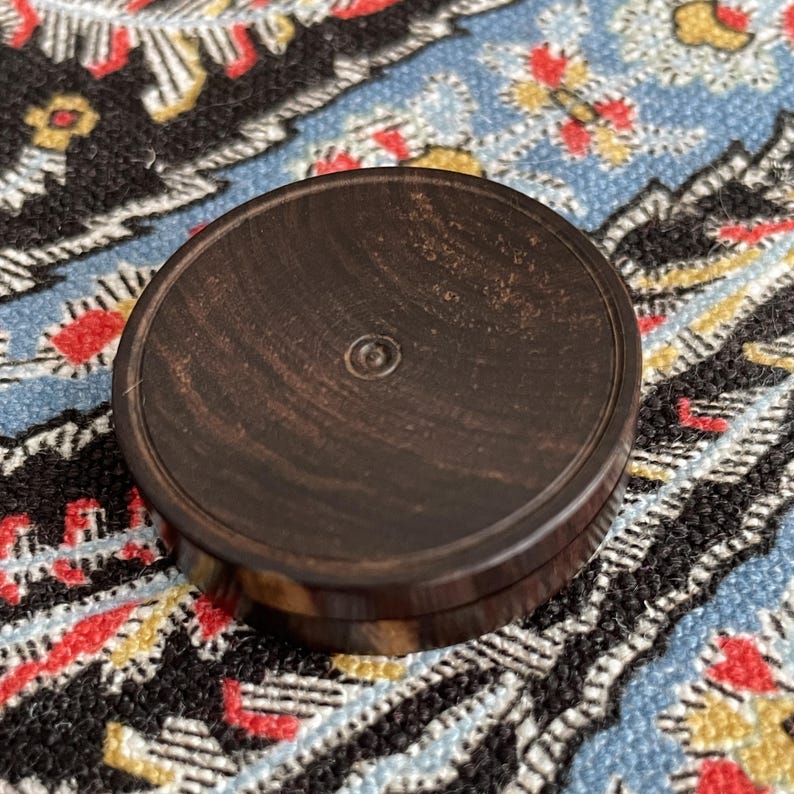 May include: A round, dark brown wooden container with a lid. The lid has a small, circular indentation in the center. The wood grain is visible on the surface. The container is sitting on a patterned fabric.