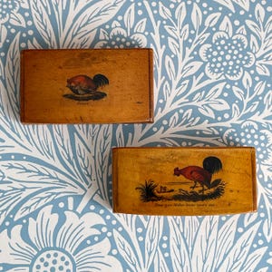 May include: Two rectangular wooden boxes with rooster illustrations. One box has a rooster illustration with the word "Orules" below it. The other box has a rooster illustration with the text "Does your Mother know you're out."