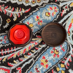 May include: A red wax seal with a laurel wreath design inside a dark wooden case. The lid of the case is open, revealing the seal. The seal is octagonal. The background is a patterned fabric with blue, black, red, and yellow colors.