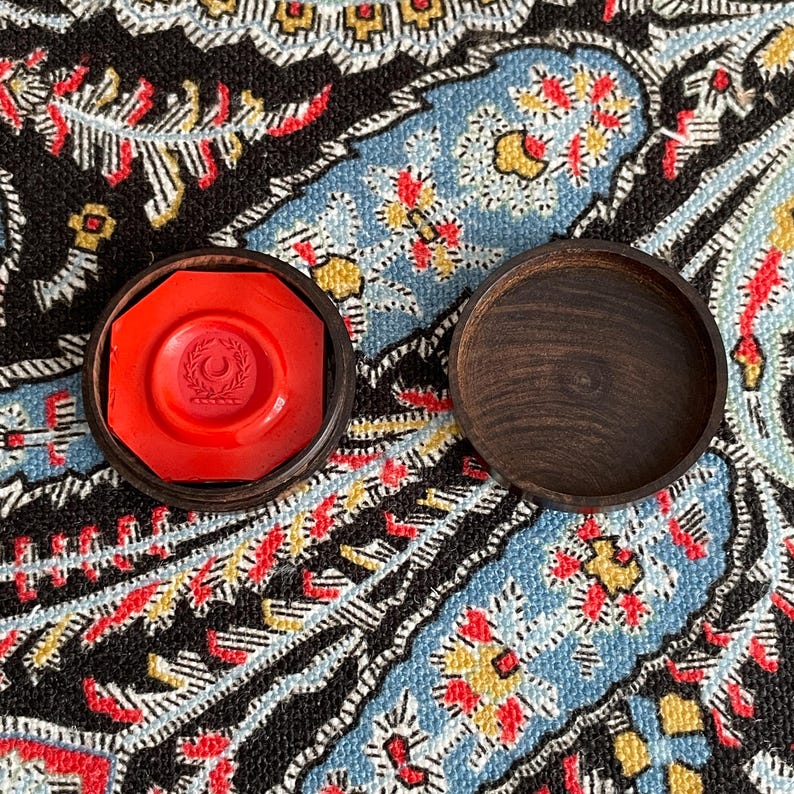 May include: A red wax seal with a laurel wreath design inside a dark wooden case. The lid of the case is open, revealing the seal. The seal is octagonal. The background is a patterned fabric with blue, black, red, and yellow colors.