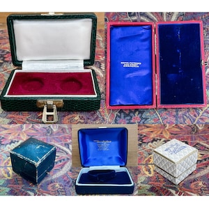 May include: Four different empty jewelry boxes. The first box is green with a red velvet interior and a latch. The second box is red with a blue velvet interior. The third box is blue with a blue velvet interior. The fourth box is white with a gold pattern and blue text.