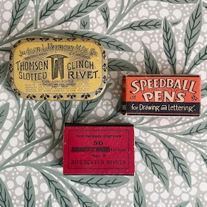 May include: Three vintage boxes on a patterned background. The top box is yellow with black text, reading "Thomson Slotted Clinch Rivet." A smaller orange box says "Speedball Pens." A red box reads "Bifurcated Rivets."