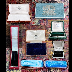 May include: Assortment of vintage jewelry boxes in various colors and styles. The boxes are made of wood and have velvet interiors. Some of the boxes have labels with the names of jewelers.