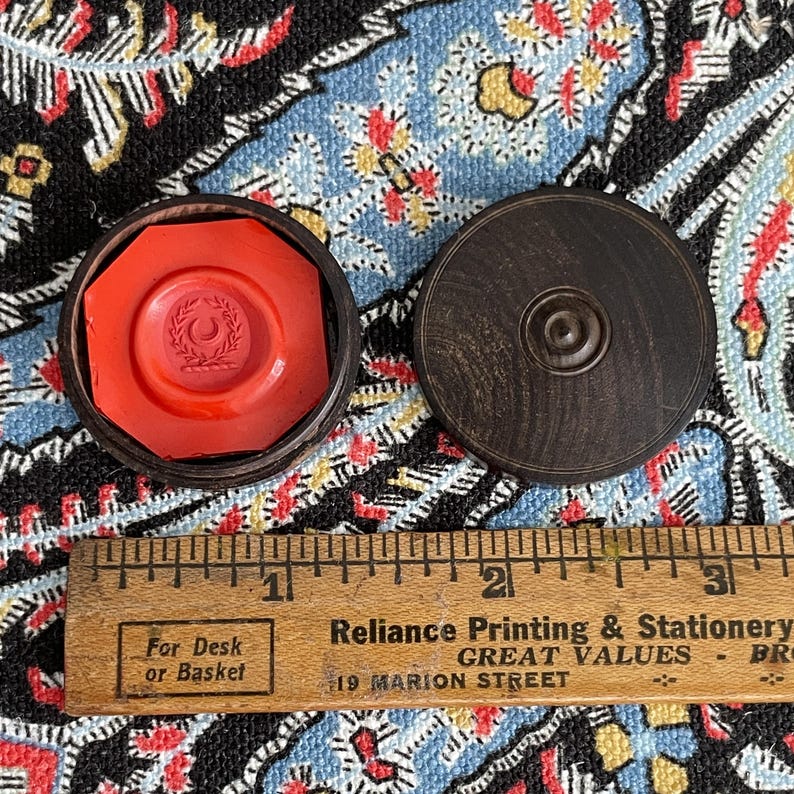 May include: An open, dark wooden box with a bright red wax seal inside. The seal has a design of a crescent moon and leaves. A wooden ruler with inch markings is in the foreground.