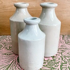May include: Three white ceramic bottles of varying sizes. The bottles have a smooth, glazed finish and a rounded, cylindrical shape. The bottles are arranged on a pink and green patterned surface.