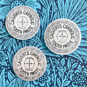 May include: Three white round tins with black lettering. Each tin has the text "Calvert's Carbolic Tooth Paste" and "Trade Mark" with a compass design in the center. The tins are on a blue and black floral patterned background.