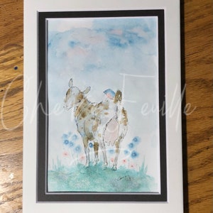 May include: Watercolor painting of a goat with a bluebird perched on its back, framed in white. The goat is brown and white, standing in a field of blue and pink flowers under a blue sky. The artwork is signed.