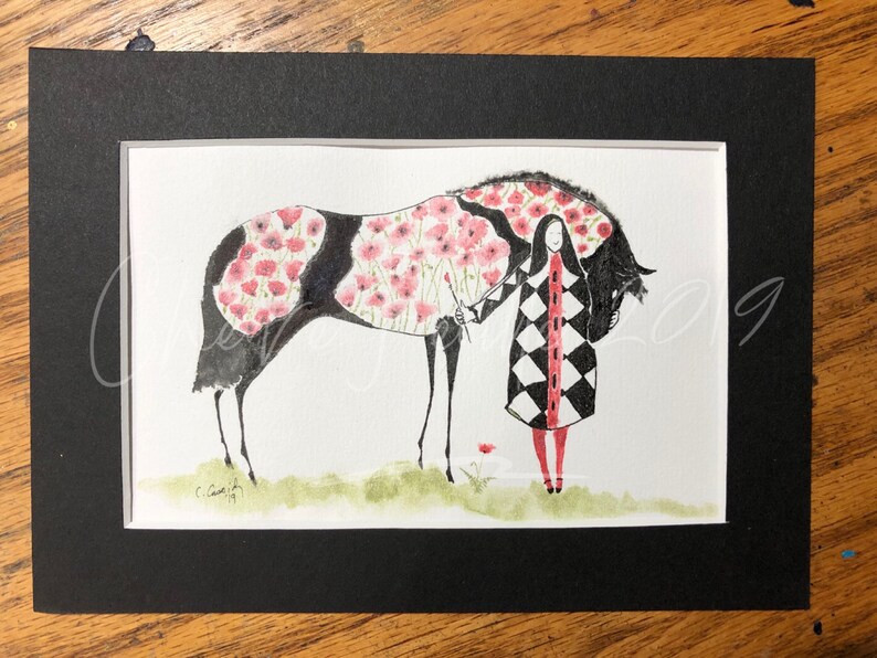 May include: A watercolor painting of a black horse with a pink floral pattern on its body. A woman in a black and white checkered coat stands next to the horse. The painting is framed in black.