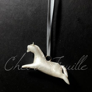 May include: A white, leaping horse ornament with a pearlescent finish, suspended by a gray ribbon. The horse is detailed with a small horn and flowing mane. The background is black.