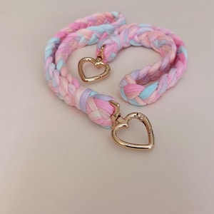 May include: Pastel-coloured braided lanyard in pink, blue, and lavender. The lanyard has gold-coloured heart-shaped clasps at each end. The braided design is made of soft fabric, creating a whimsical and stylish accessory.