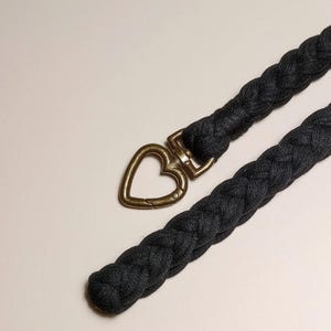 May include: A black braided fabric strap with a heart-shaped metal clasp. The clasp is gold-toned.