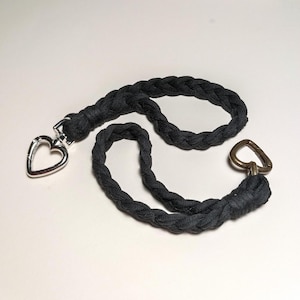 May include: A black braided fabric cord with a silver heart-shaped clasp on one end and a gold heart-shaped clasp on the other end.