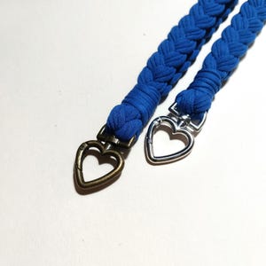 May include: Two blue braided straps with heart-shaped clasps. One clasp is silver, and the other is bronze. The straps are made of a soft, woven material. The background is white.