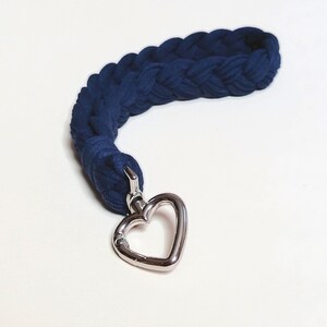 Pode incluir: Uma pulseira trançada azul marinho com um fecho prateado em forma de coração. A pulseira é feita de um material espesso e macio e foi projetada para ser usada no pulso. O fecho de coração adiciona um toque de elegância.