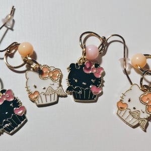 May include: Four gold-toned dangle earrings. Each earring features a charm shaped like a cupcake with a Hello Kitty design. The charms are black and white with pink accents. Each earring has a pink and peach-colored bead.